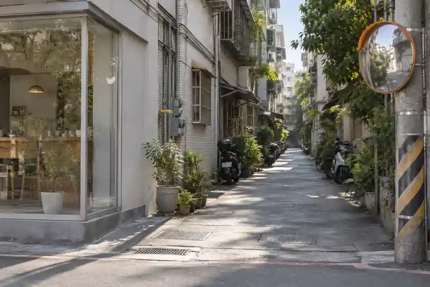 大馬路轉進巷子的路口：電線桿上掛著橘色框的圓形凸面鏡，巷子往裡延伸，兩旁停著機車